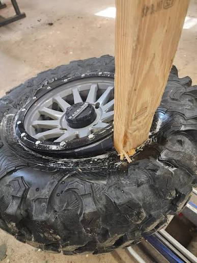 A 2x4 wooden plank driven into the bead area of an off-road tire on a beadlock-style wheel as an improvised bead-breaking method